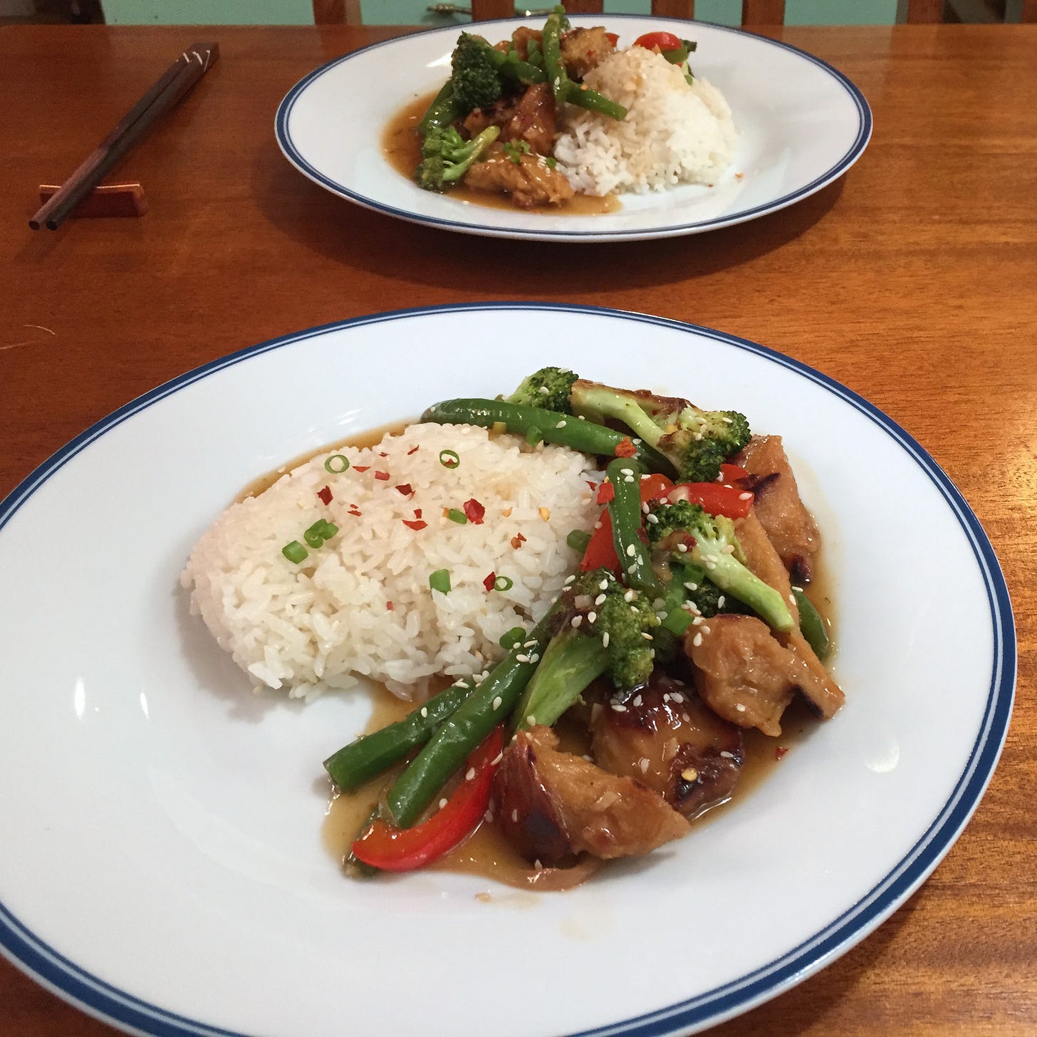 Two plates on a wooden table next to chopsticks on wooden rests. On the plates is scoops of rice next to green beans, broccoli, red pepper, and seitan in orange sauce.