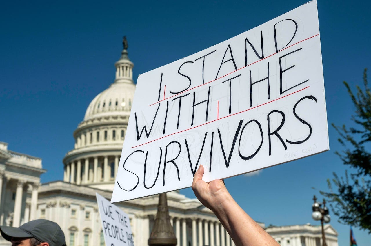 A white sign with black lettering reading I stand with the survivors, is held up against a backdrop of the Capitol Building
