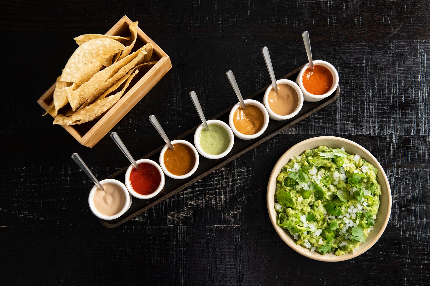 tostadas in a wooden box next to a selection of seven salsas in white ramekins and a bowl of guacamole, all atop a black wooden tabletop