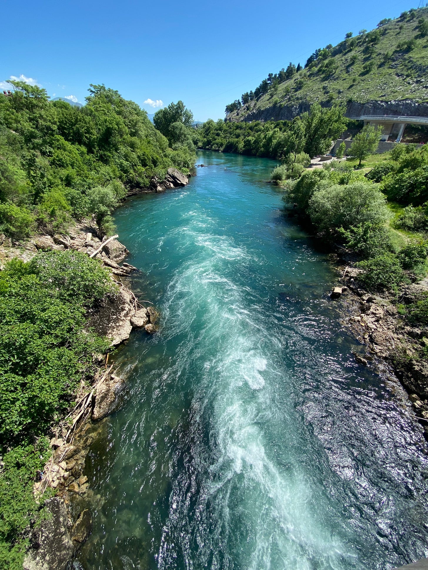  An elevated, high-angle view looking down a vibrant, turquoise river as it flows through a lush, green canyon under a clear blue sky. The water is bright teal and transitions into white, churning rapids in the foreground, creating a sense of powerful movement. Dense green trees and shrubs line both rocky banks, and a steep, sunlit hill rises on the right side. In the upper right background, a concrete bridge structure is visible, partially tucked into the hillside. The overall scene is bright, natural, and serene.