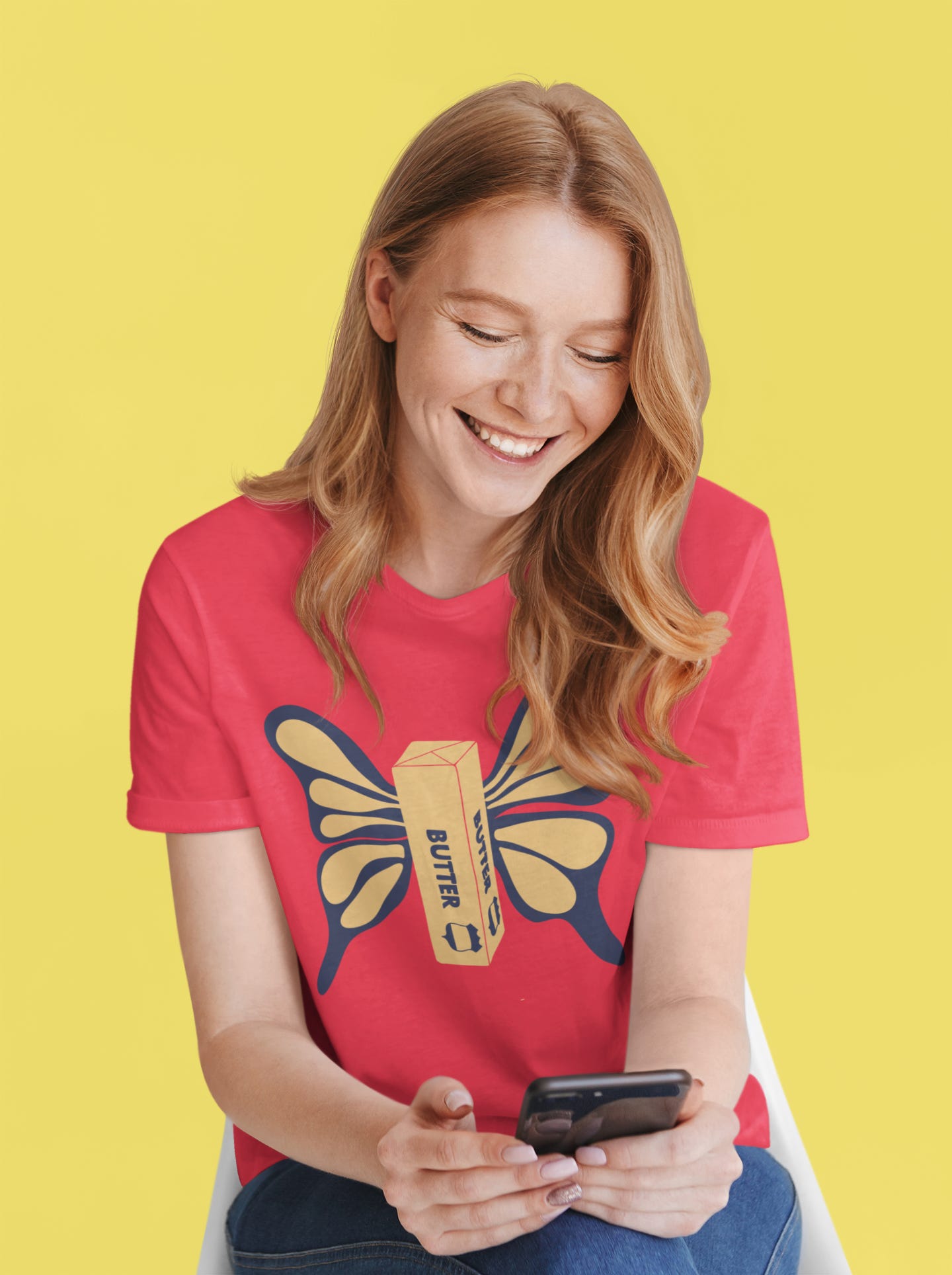 Stock photo of a woman reading something on her phone wearing a red t-shirt with a butterly graphic on it, where the insect's body is a sticke of butter.