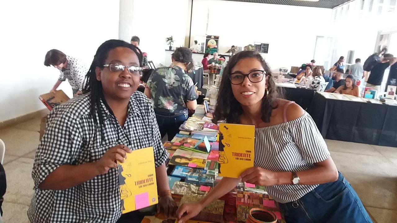 Foto horizontal. Em primeiro plano, estou no lado direito da imagem, inclinada sobre uma mesa preenchida com vários livros. No lado esquerdo, alinhada comigo, está a escritora Cidinha da Silva, uma mulher negra com óculos retangulares e cabelo trançado, vestindo uma camisa quadriculada preta e branca. Cada uma de nós segura um exemplar do livro "Cada tridente em seu lugar", de Cidinha. Atrás de nós, se vê o fim da mesa e outras pessoas, que olham livros, mexem em caixas ou estão sentadas em cadeiras no fundo.