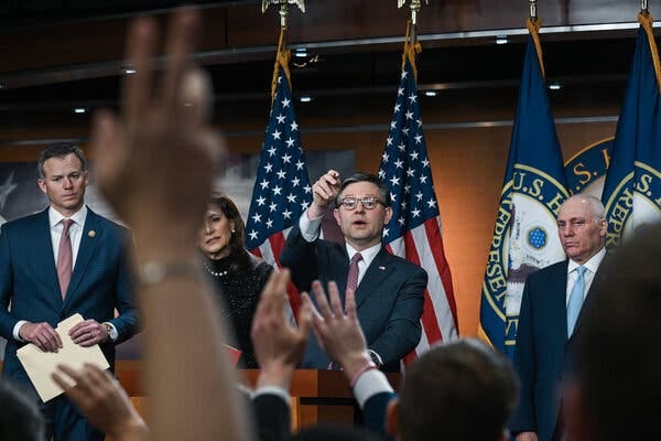 Speaker Mike Johnson, in a dark suit and a red tie, raises his hand to call on a reporter during a news conference at the Capitol.