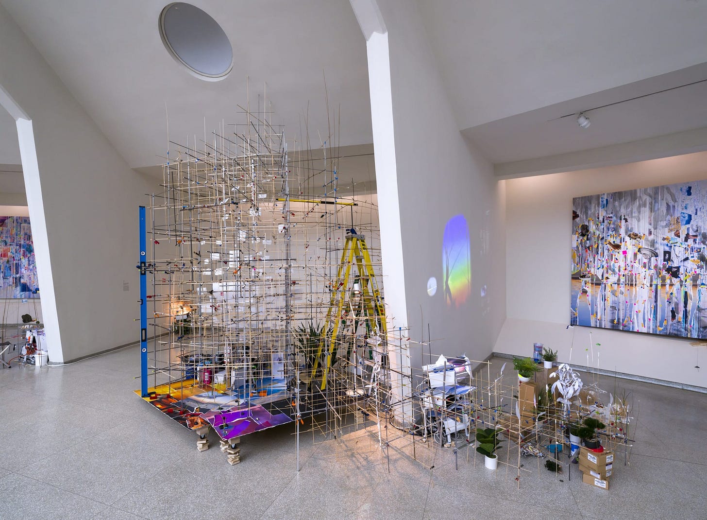 A view of two bays of the Guggenheim's spiral with installations by Sarah Sze. A tall scaffolding of thin sticks supports torn pieces of paper which serve as screens for projected images. A yellow stepladder is visible behind the structure. In the corner of the bay you can see the light from a projector. In the next bay the scaffolding continues as a low structure. Behind it is a painting of complex collaged layers. Several potted plants and cardboard boxes are also visible in the arrangment.