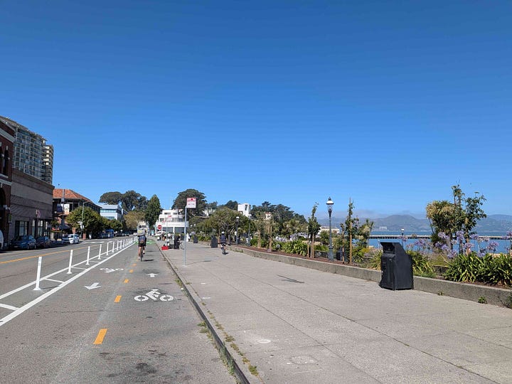 1: Bikeway along the waterfront, the Marin headlands is seen in the distance, 2:Bikeway in front of Ghirardelli Square. Bikeway is painted green. A man is cross the street.