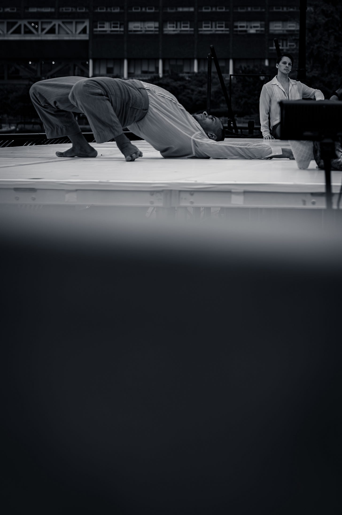 A dancer lies arched on their back on stage, while another performer looks on from the side of the platform. A dancer lies arched on their back on stage, while another performer looks on from the side of the platform.