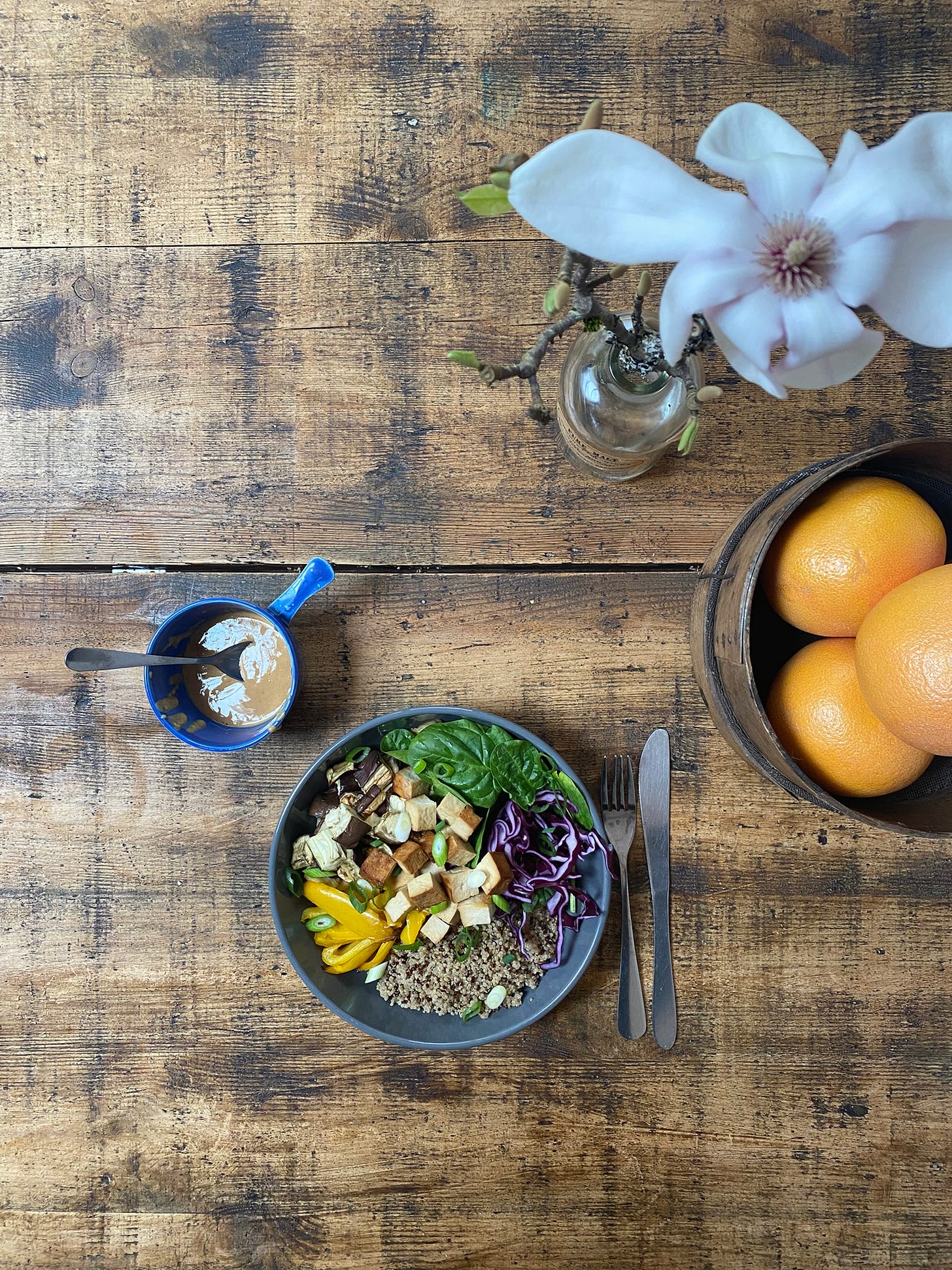A wooden table top set with a branch of blossoming flowers, a wooden bowl containing pink grapefruit, a bowl of mixed foods containing spinach, cubes of tofu, sliced red cabbage, yellow peppers, and grain. Next to the bowl, a blue cup contains a beige glossy sauce.