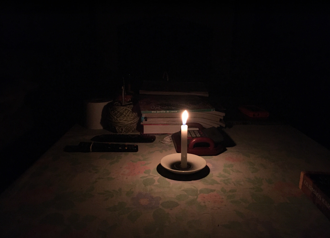 A single lit candle in a white holder glows in darkness, illuminating a desk scattered with books, a ball of twine, pens, and a flashlight. The candle's warm light creates a small circle of visibility against the surrounding blackness, suggesting a power outage.