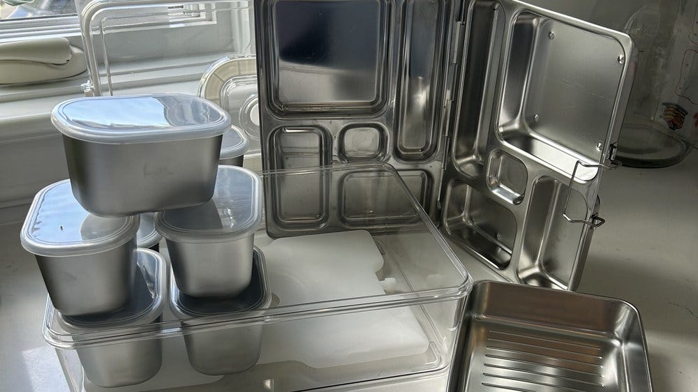 A stainless PlanetBox lunch box, a stainless snack container box, and stacked stainless fridge containers arranged on a kitchen counter. A stainless PlanetBox lunch box, a stainless snack container box, and stacked stainless fridge containers arranged on a kitchen counter.