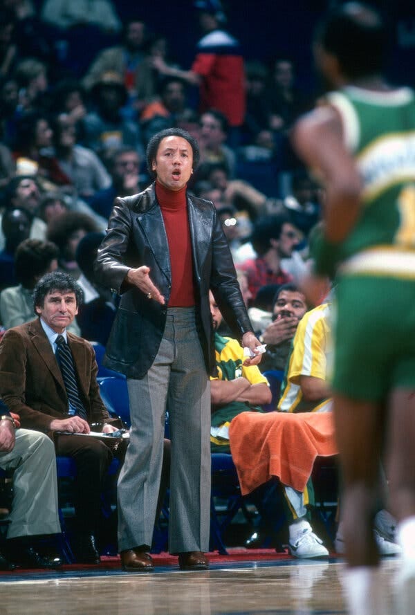 A man in a leather jacket stands on a sideline of a basketball court. A man in a leather jacket stands on a sideline of a basketball court.