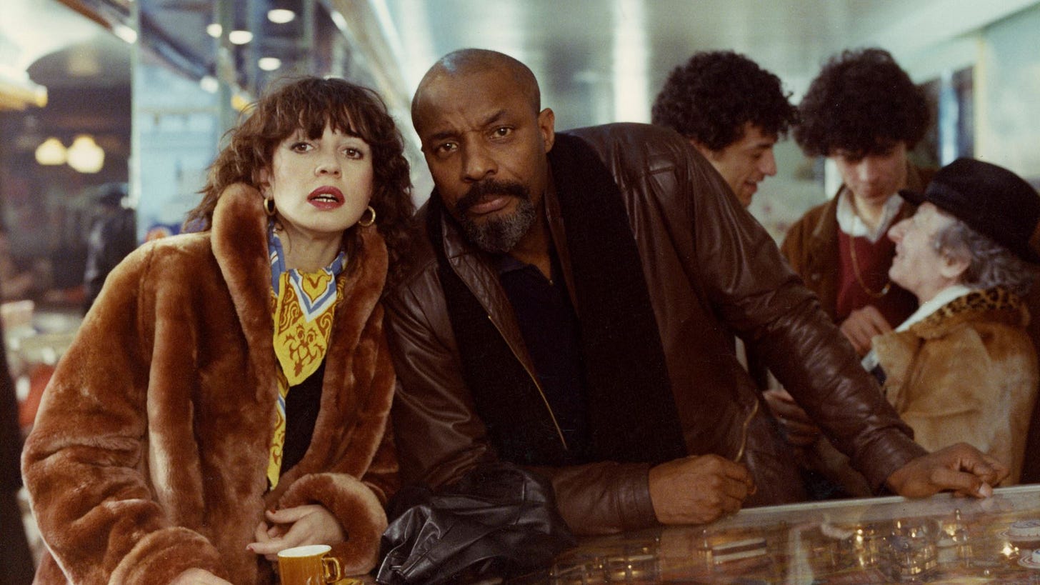Juliet Berto as Anita, a young to middle aged white woman with red lipstick shaggy hair and a huge fur coat, looking off into the distance and standing at a pinball machine leaning into Robert Liensol as Jocko, a middle aged Black man looking sternly into the camera, wearing a brown leather jacket Juliet Berto as Anita, a young to middle aged white woman with red lipstick shaggy hair and a huge fur coat, looking off into the distance and standing at a pinball machine leaning into Robert Liensol as Jocko, a middle aged Black man looking sternly into the camera, wearing a brown leather jacket