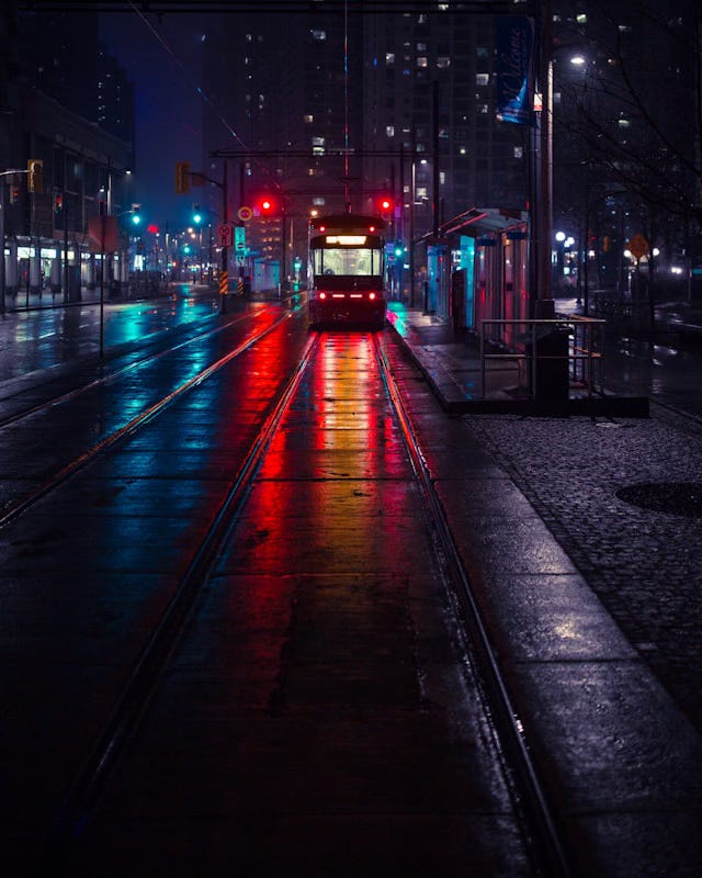 A dark, wet night in a city. A street car sits at a station at the end of a track, with the street stretching parallel to the left and a cobblestone walk to the right. Tall buildings reach to the sky on the left and in the distance.