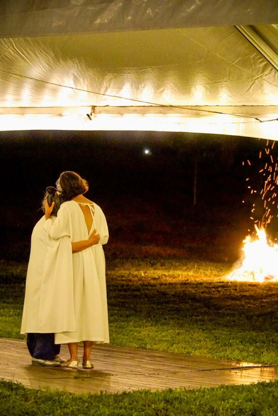 Two women stand, holding one another lovingly, while turned away from the camera gazing at a robust bonfire against a dark night sky. They stand underneath a lighted tent on top of a dance floor placed on grass. Two women stand, holding one another lovingly, while turned away from the camera gazing at a robust bonfire against a dark night sky. They stand underneath a lighted tent on top of a dance floor placed on grass.