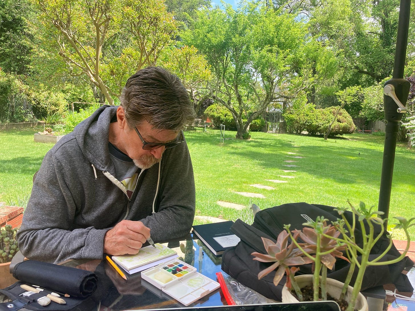 A man paints a watercolor in a lush, green and sunny backyard