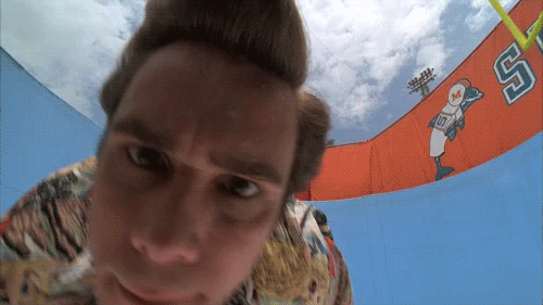 Screenshot from the movie Ace Ventura showing Jim Carrey's character with an exaggerated surprised/excited facial expression, shot from a low angle with a colorful background.