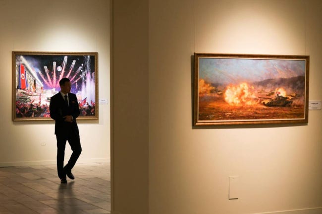 Un homme déambule dans une galerie. Un tableau montre un tank en feu, un autre des bannières aux couleurs de la Corée du Nord décorant une avenue