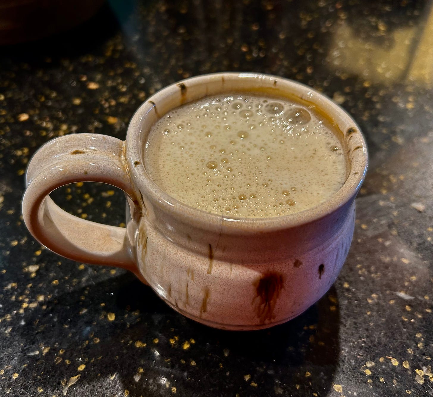 A tan with brown flecks ceramic coffee mug with foamy foam on top of its coffee. It sits on a black with gold-fleck stone countertop