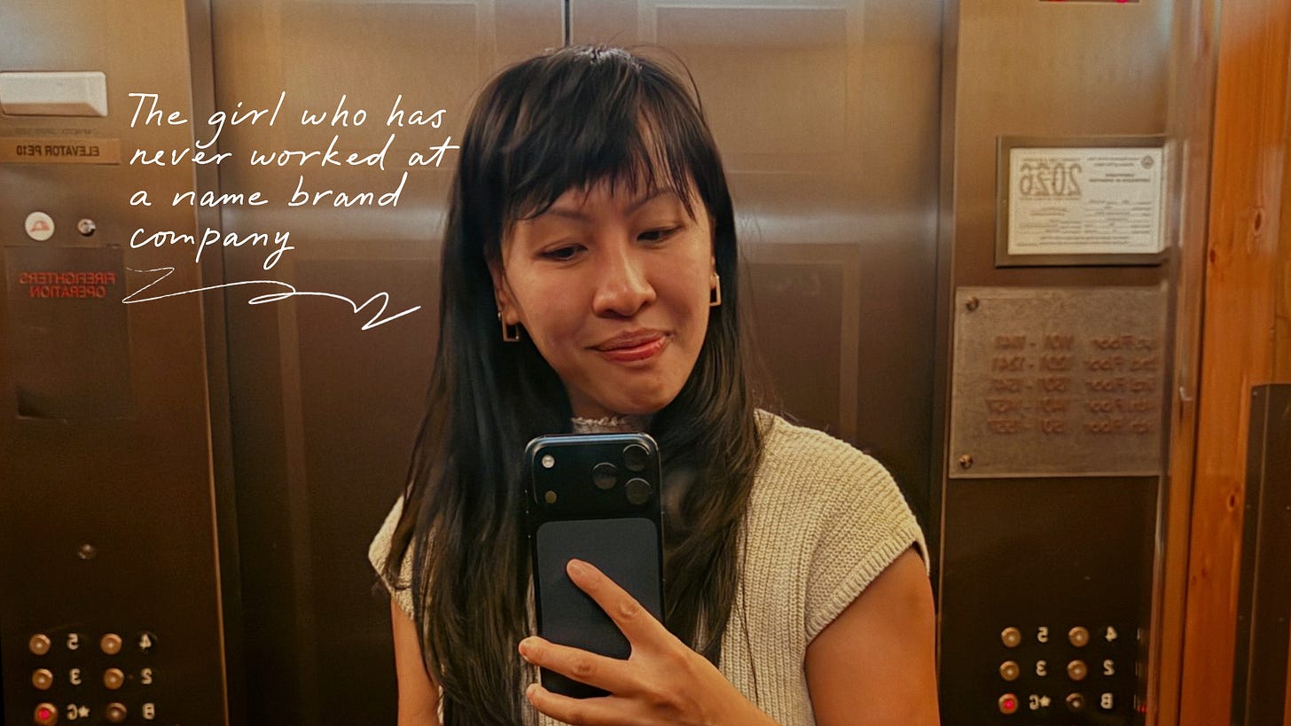 Mirror selfie of a woman standing in an elevator holding her phone. Handwritten text beside her reads, “The girl who has never worked at a name brand company,” with an arrow pointing toward her.