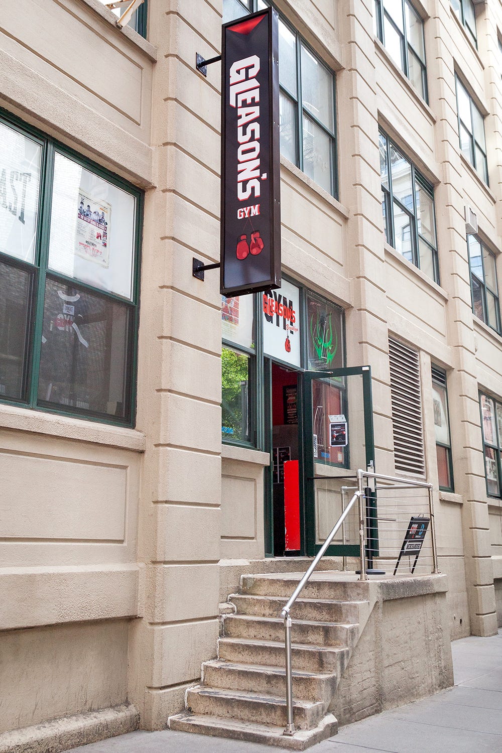 The entrance to Gleason's Gym at 130 Water Street in DUMBO, Brooklyn, with a vertical red and black Gleason's Gym sign above concrete steps and a metal railing