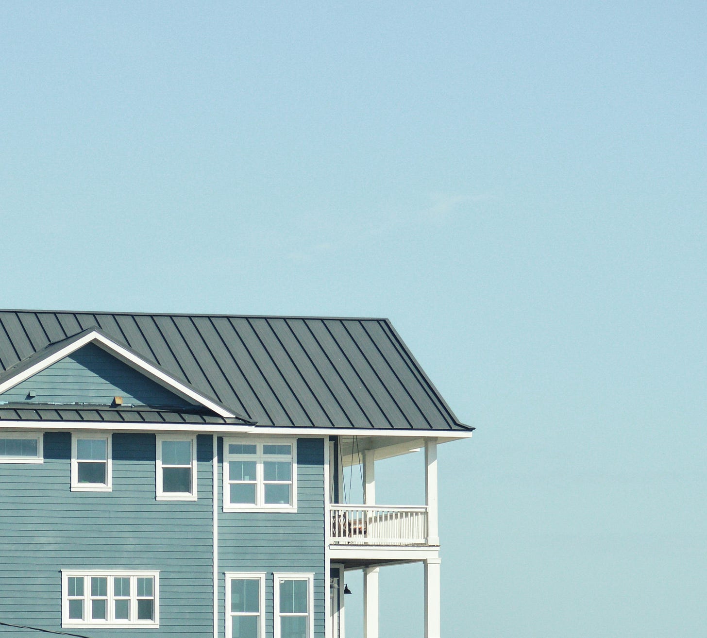 Side view of a house with blue siding and a grey metal roof. The house has multiple windows with white frames and a sunlit balcony. Side view of a house with blue siding and a grey metal roof. The house has multiple windows with white frames and a sunlit balcony.