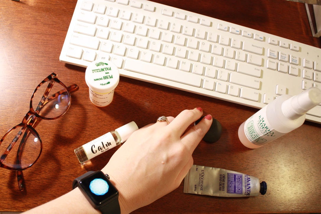 A hand using a mouse wearing an Apple Watch that is running the breathing app. The desk has glasses, a pill bottle, a "calm" sent roller, hand lotion, and rose face spray.