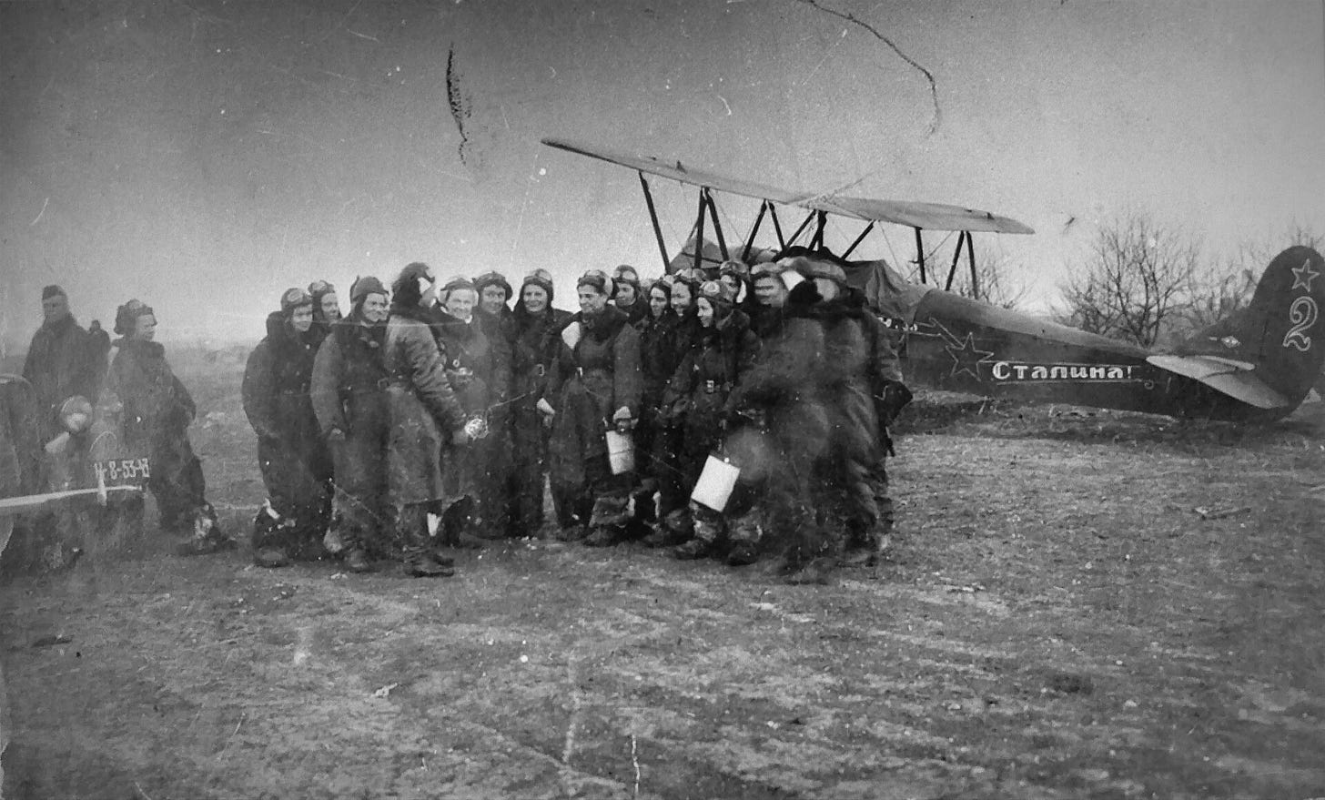 About twenty Soviet women in flight suits and leather flying helmets stand together on a muddy airfield in front of a Po-2 biplane with a slogan painted on its fuselage. About twenty Soviet women in flight suits and leather flying helmets stand together on a muddy airfield in front of a Po-2 biplane with a slogan painted on its fuselage.