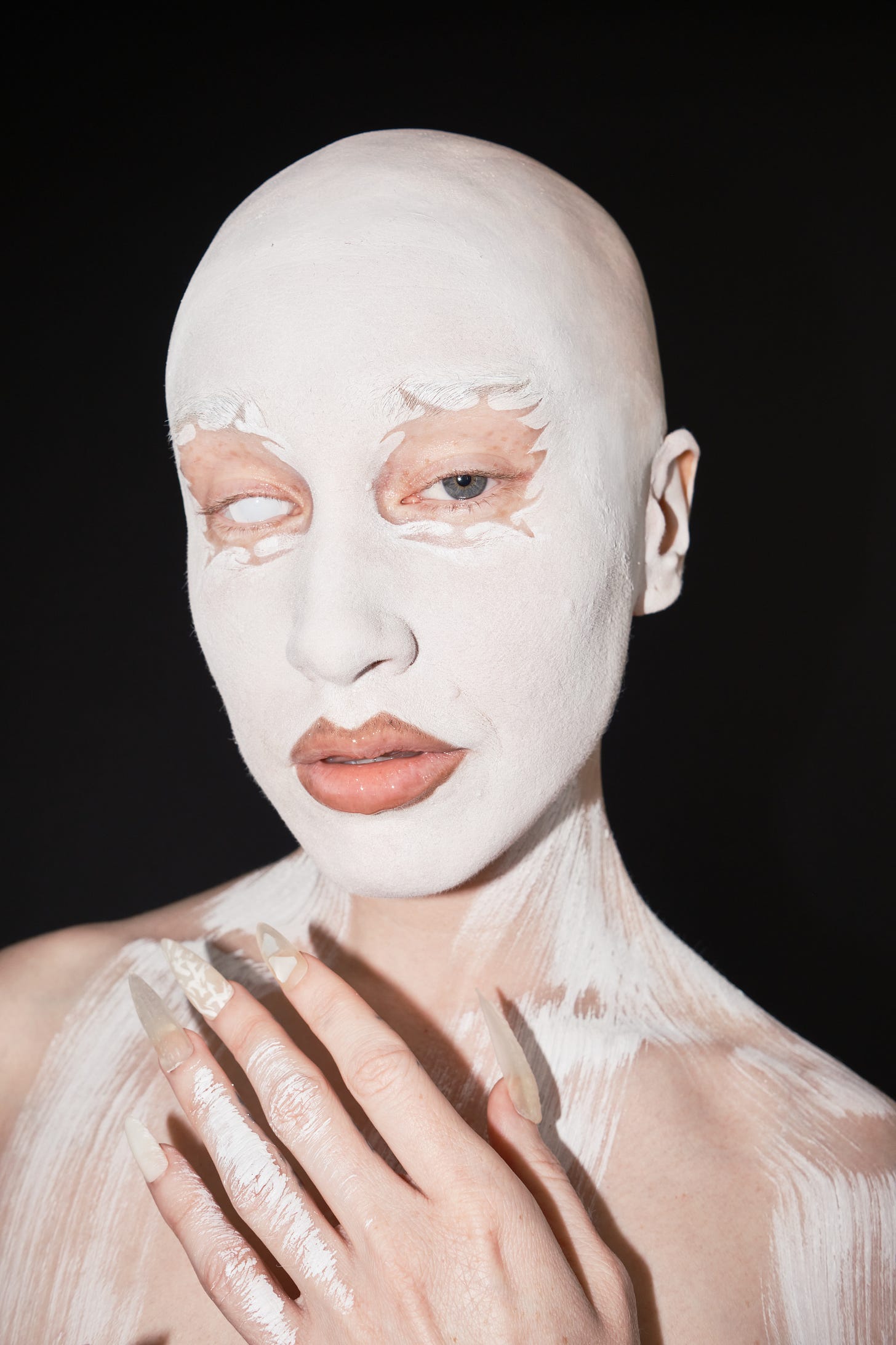 a white person wearing white face paint (except around their eyes) looks at the camera. One eye is completely white, the other is green.