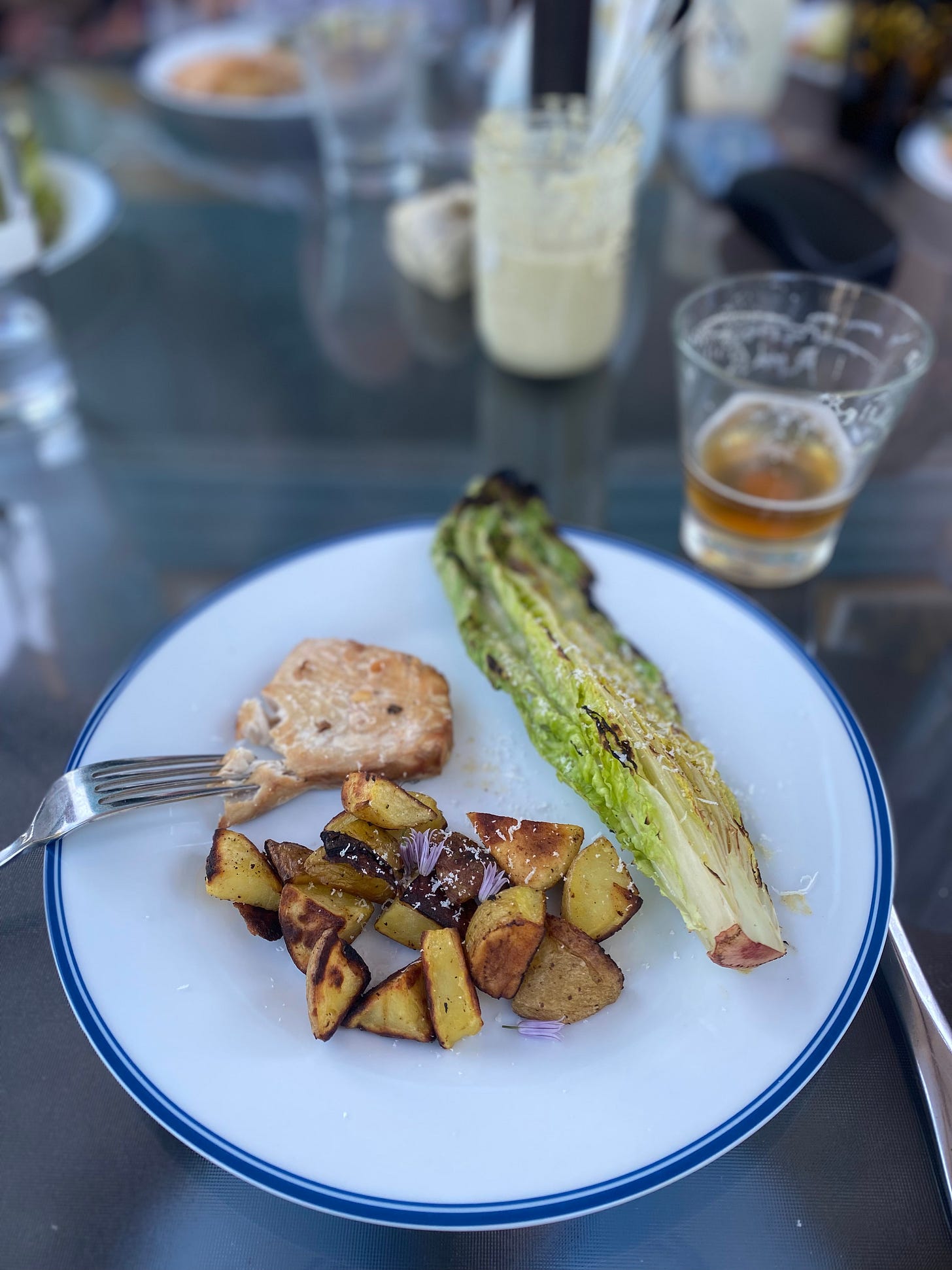A dinner plate on an outdoor glass table with salmon, crispy potato pieces, and half a romaine heart, all dusted with parmesan and chive blossoms.