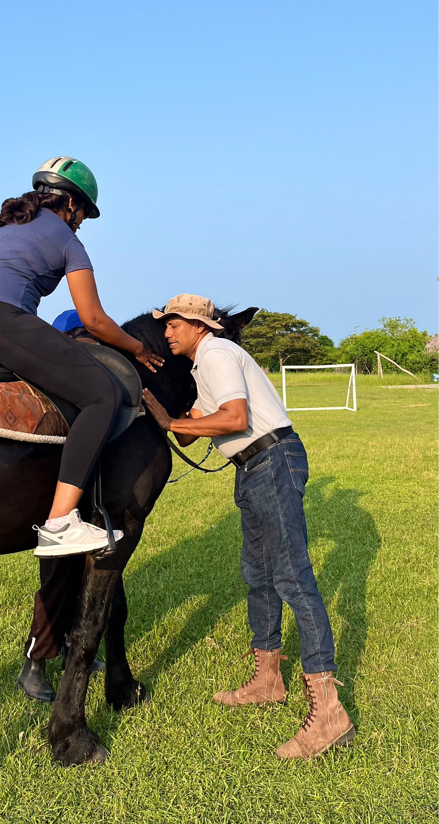 Alfie stands beside a horse, calmly petting and reassuring it, while teaching a rider who also reaches out to comfort the horse’s neck. The moment captures leadership, trust, and communication in a beautiful natural setting.
