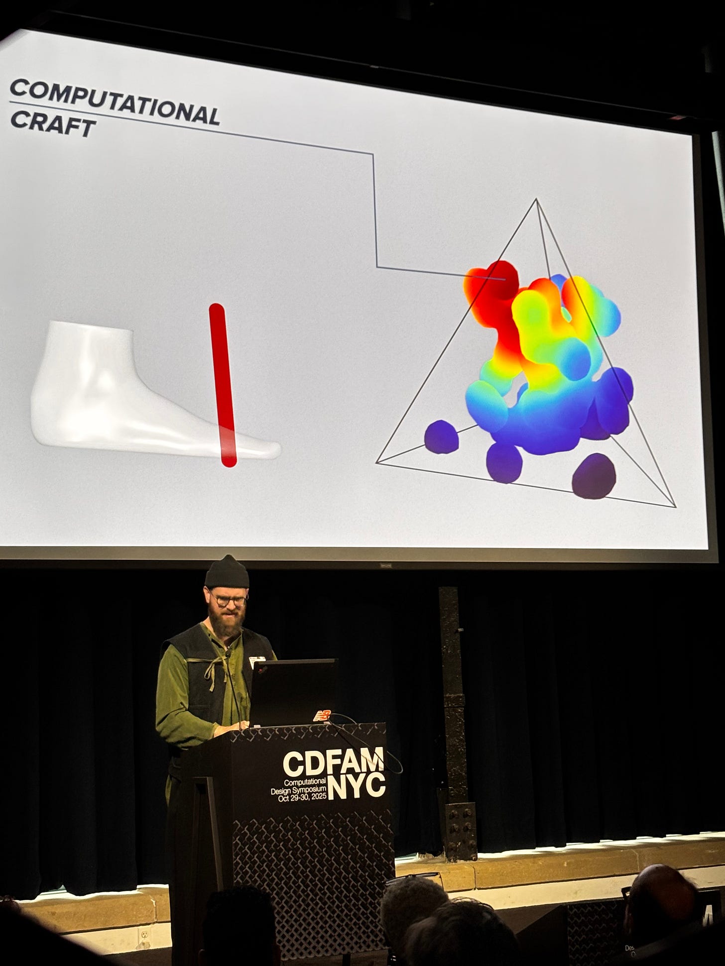 A presenter stands at a podium labeled 'CDFAM NYC', displaying a visual presentation on computational design. The slide features a 3D model of a footwear design alongside colorful, fluid-like shapes in a triangular layout. A presenter stands at a podium labeled 'CDFAM NYC', displaying a visual presentation on computational design. The slide features a 3D model of a footwear design alongside colorful, fluid-like shapes in a triangular layout.
