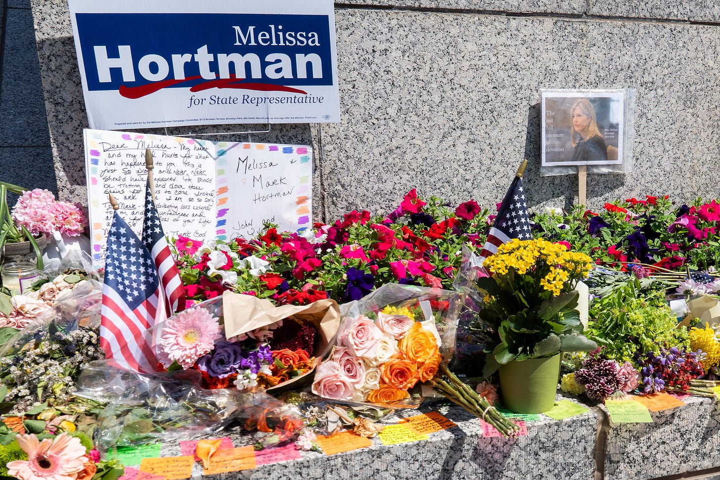 American flags, flowers, cards, and post-it messages along the edge of larger stone monument. In the top left corner, a campaign sign reads, "Melissa Hortman for State Representative."