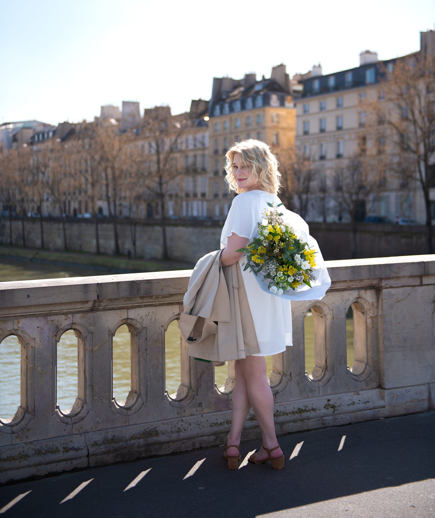 Karen Bussen stands in a white dress with a large bouquet of white and yellow flowers on a bridge in Paris, France