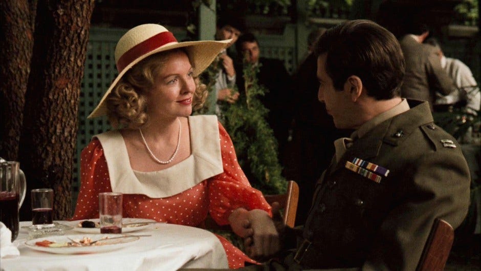 Diane Keaton as Kay Adams and Al Pacino as Michael Corleone sit at a table. She is wearing a sun hat, pearls, and a red dress with white spots. He is wearing a WWII uniform. Diane Keaton as Kay Adams and Al Pacino as Michael Corleone sit at a table. She is wearing a sun hat, pearls, and a red dress with white spots. He is wearing a WWII uniform.