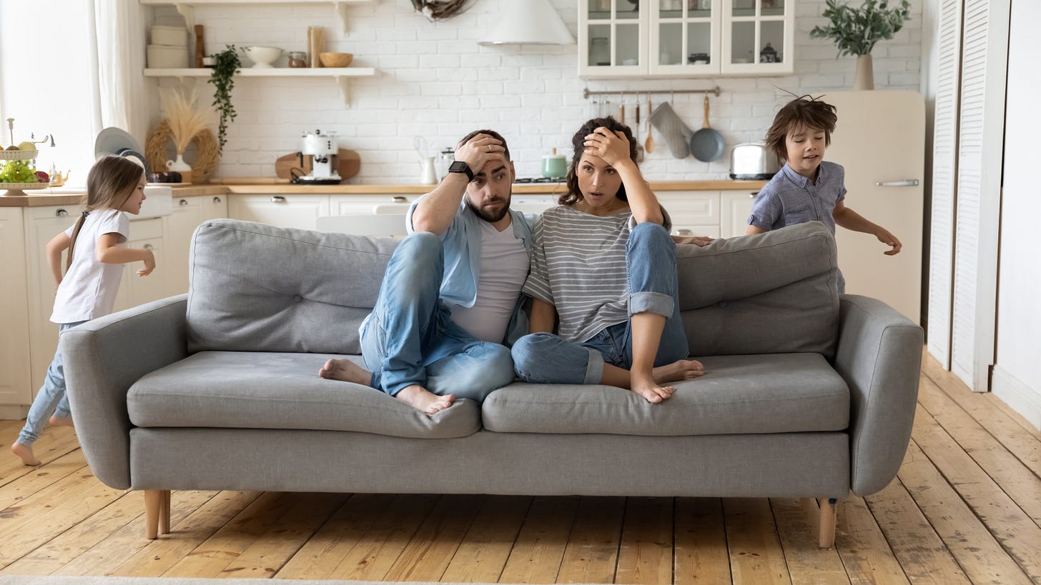 Two overwhelmed parents while children run around them