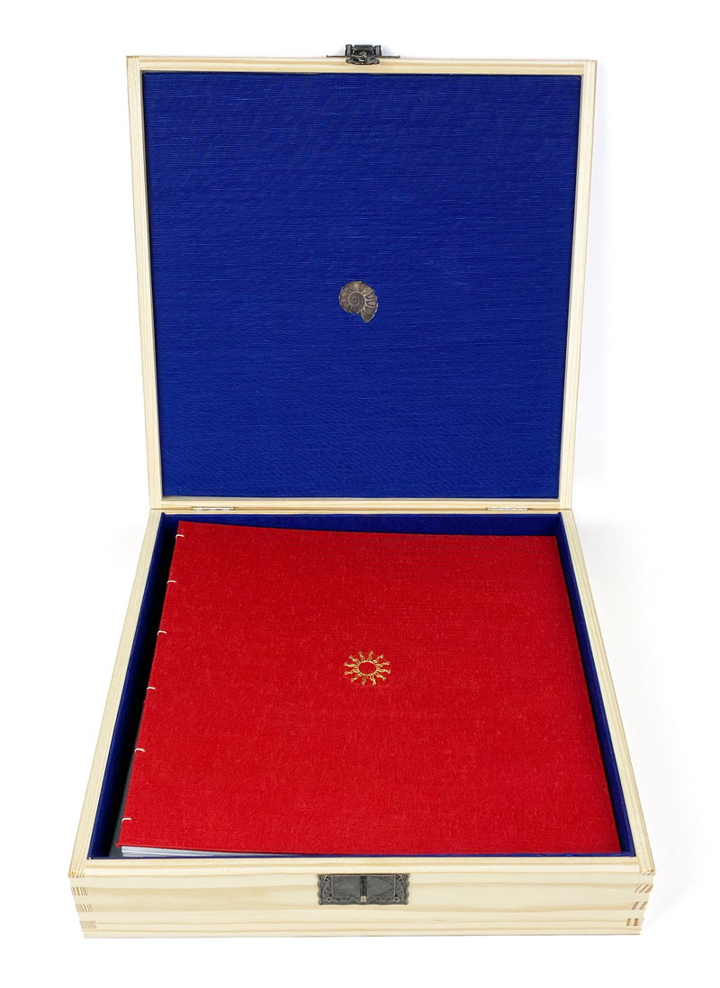 Square wooden box, open seen frontally, showing blue silk lining inside the lid and containing a red book 