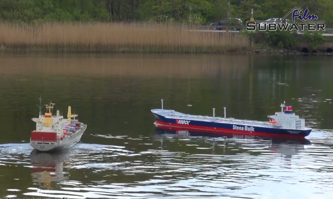 two radio controlled model ships in a calm pond, with trees, plants, and full-sized cars on a road along the shore clearly visible in the background. 