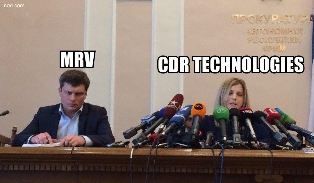Press conference photo. A man at the table labelled "MRV" sits with no microphones; beside him a woman labelled "CDR technologies" faces a thicket of microphones.