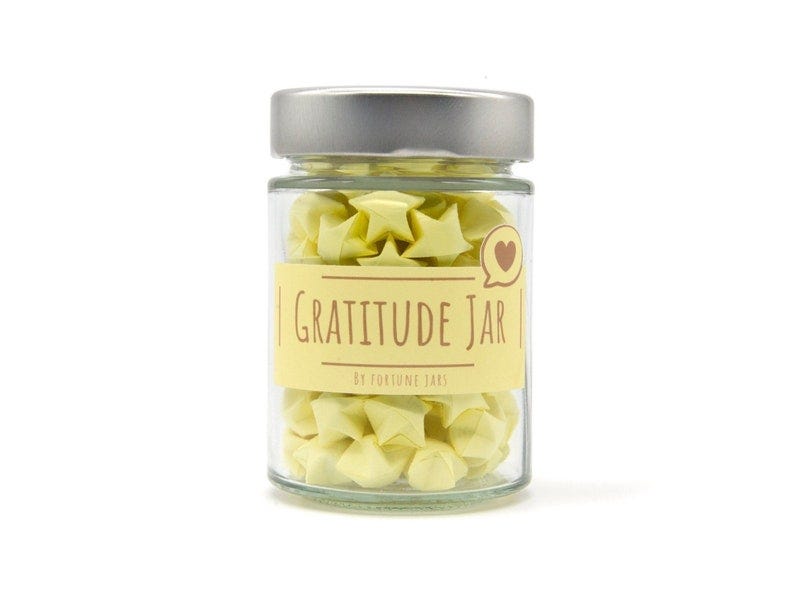 May include: A small glass jar with a silver lid contains pale yellow origami stars. A label on the jar says 'GRATITUDE JAR' with smaller text below that reads 'BY FORTUNE JARS'. A small speech bubble with a heart icon is also on the label. May include: A small glass jar with a silver lid contains pale yellow origami stars. A label on the jar says 'GRATITUDE JAR' with smaller text below that reads 'BY FORTUNE JARS'. A small speech bubble with a heart icon is also on the label.