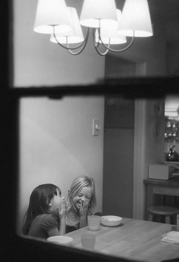 Dos niñas ríen sentadas a la mesa bajo una lámpara de techo, compartiendo un secreto. Foto en blanco y negro, vista desde fuera de la ventana.