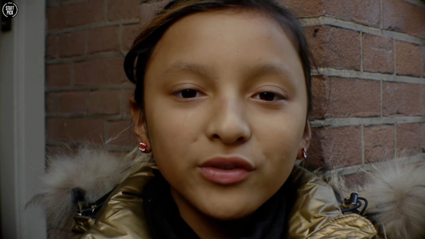 Close-up portrait of an Asian teenage girl with dark hair pulled back, wearing sparkly hoop earrings and a fur-trimmed jacket, looking directly at the camera against a brick wall.