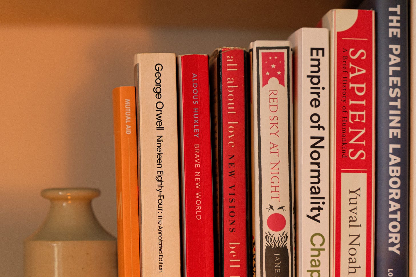 A warmly lit photo of a bookshelf illuminated by candlelight. The shelves are filled with books exploring the beauty and brutality of life stories of humanity’s past, present, and possible futures. Some are well-worn, others still waiting to be opened. The atmosphere is soft, thoughtful, and timeless.