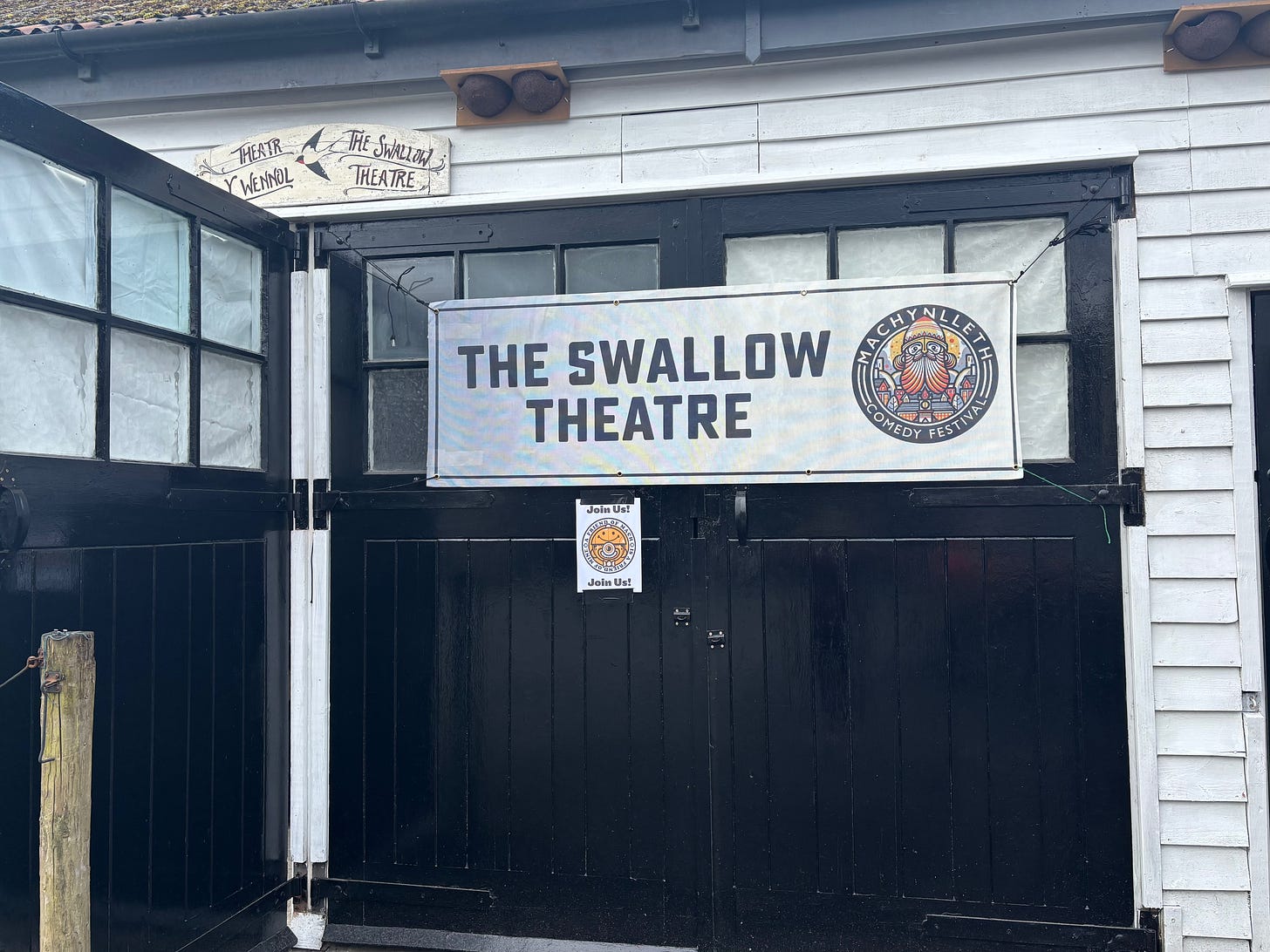 Small barn like building with white wooden slats and a black door. Poster on the door reads "The Swallow Theatre" Small barn like building with white wooden slats and a black door. Poster on the door reads "The Swallow Theatre"