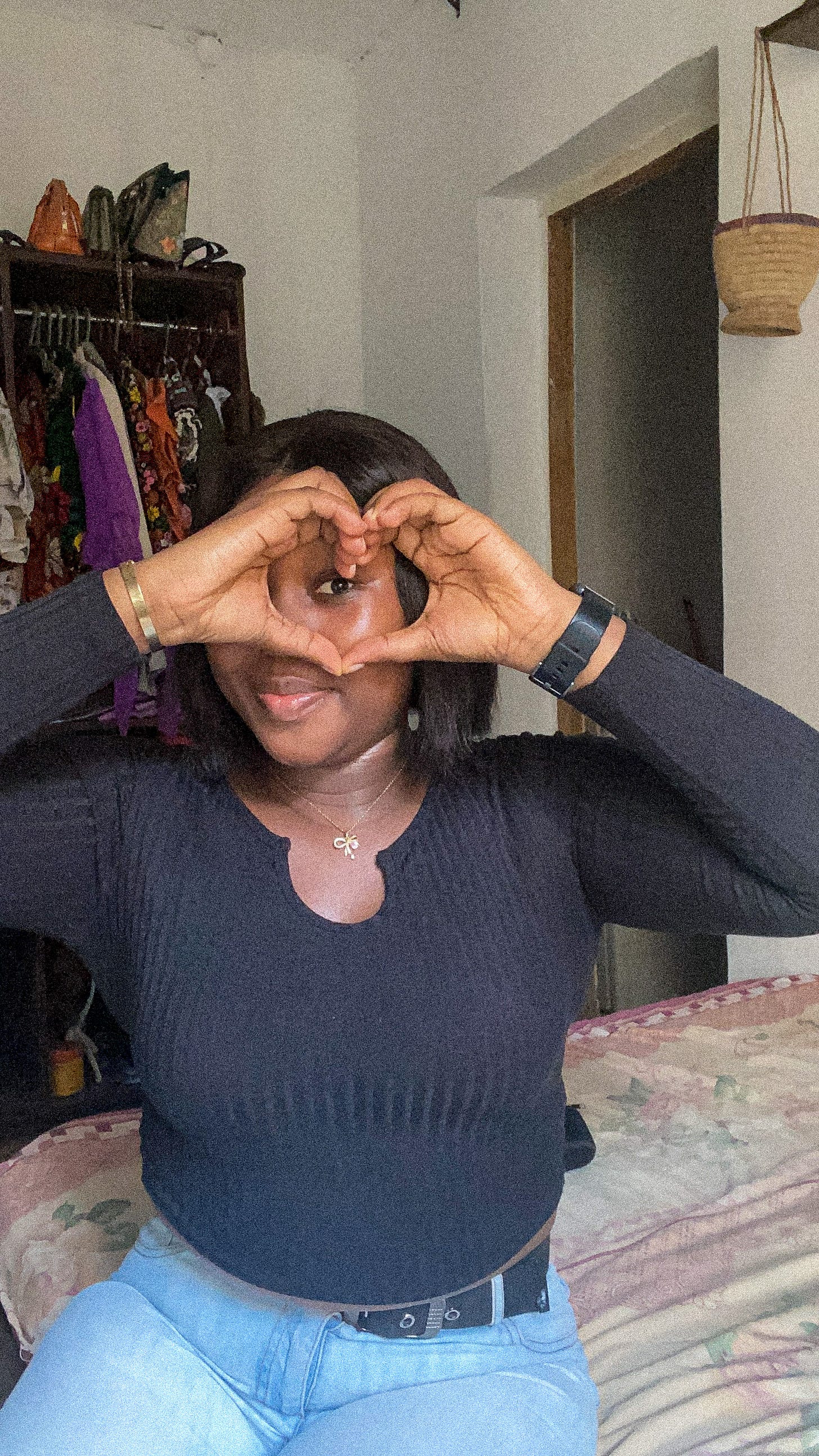 A picture of Olanike Akinyemi in black long-sleeved top and a jean trouser. She is sitting on her bed and makes a heart shape with her finger over an eye.