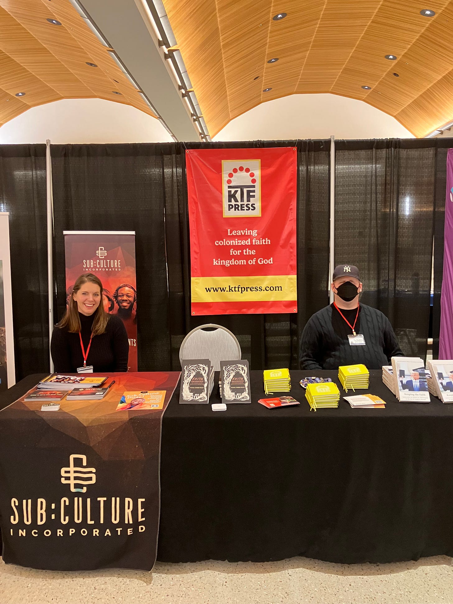 A photo of Sy standing at KTF’s booth at the Evolving Faith Conference. The booth is shared with Sub:Culture, Inc. Tamice’s nonprofit. A tall White woman is also at the booth with Sy standing on the side of the table dedicated to Sub:Culture. KTF’s books and other materials are on the table, and a large sign for KTF is hung behind Sy.