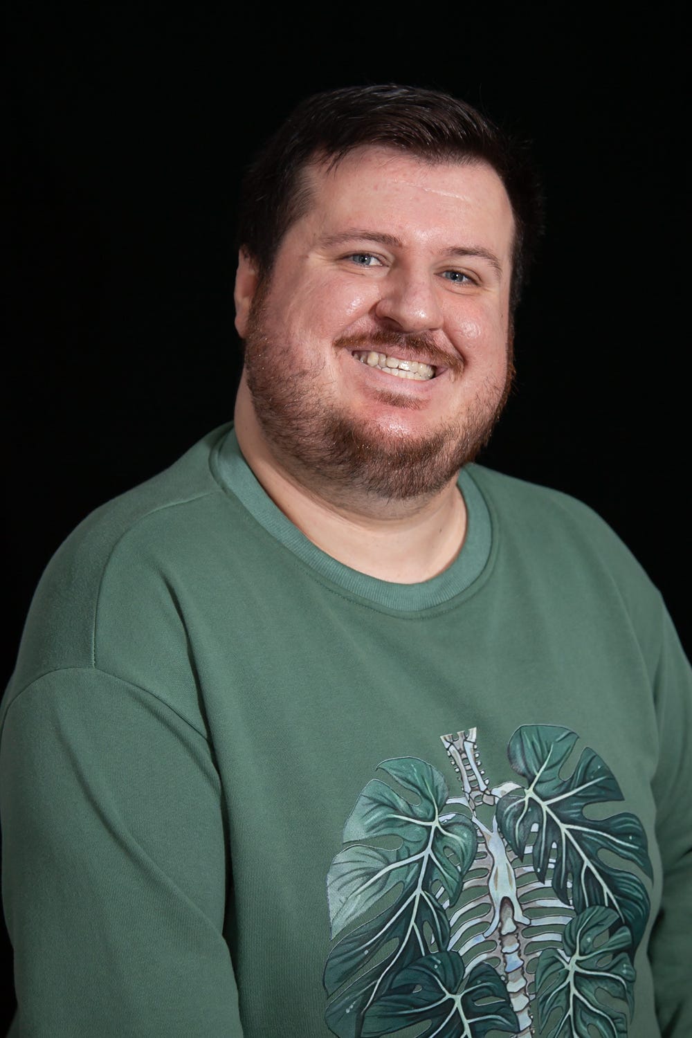 A portrait of Jordan from the mid-waist up. He is wearing a sage green sweatshirt with an image of a ribcage surrounded by leaves. He has white skin, short brown hair and blue eyes. He is looking into the camera and smiling. A portrait of Jordan from the mid-waist up. He is wearing a sage green sweatshirt with an image of a ribcage surrounded by leaves. He has white skin, short brown hair and blue eyes. He is looking into the camera and smiling.
