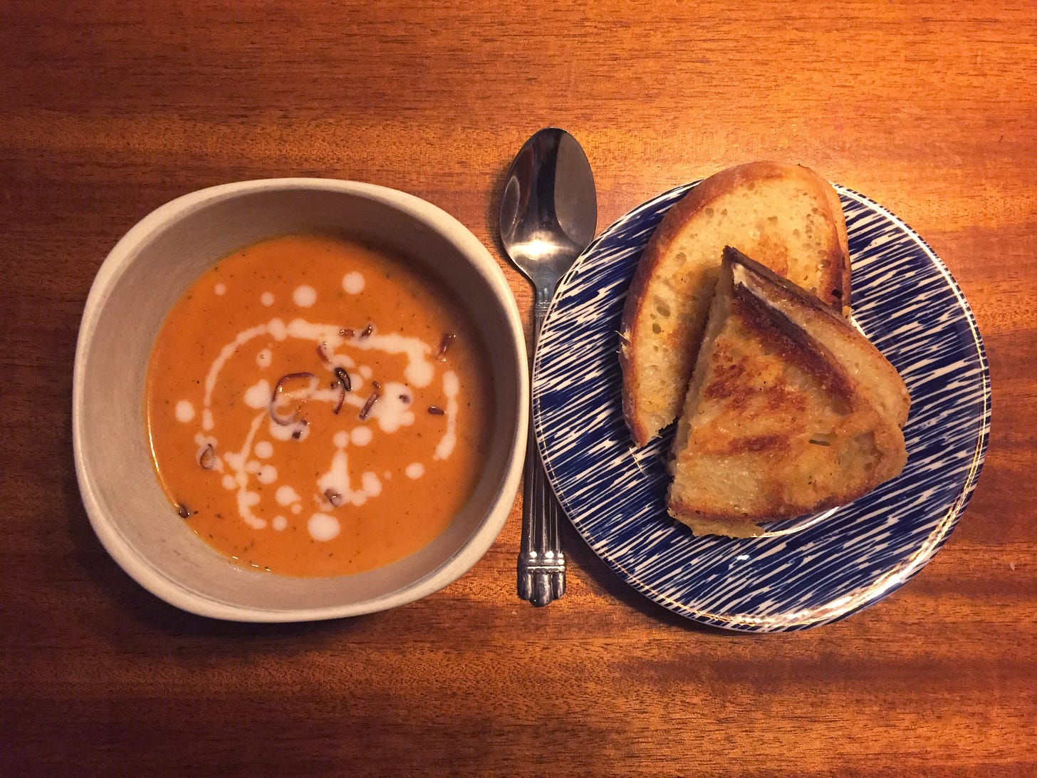 A white bowl of tomato soup with a drizzle of coconut milk and a scattering of fried shallots. Next to it, a small blue plate with halves of a grilled cheese sandwich stacked offset each other. A spoon is in between the two dishes.