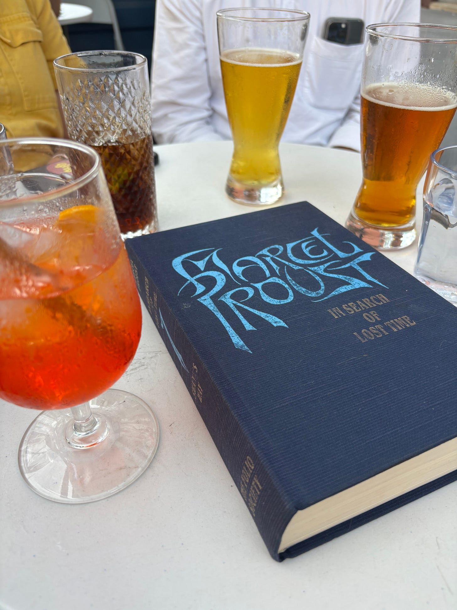 Blue hardcover of Marcel Proust's Swann's Way in the middle of a white table, surrounded by half-full drink glasses
