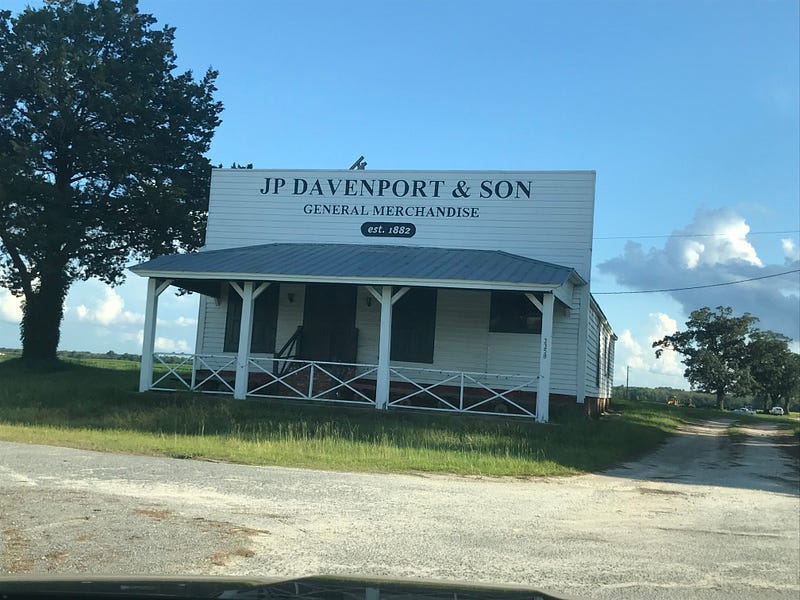 Tall white building with porch. Top of the building has “JP Davenport & Sons General Merchandise” in big navy blue letters.