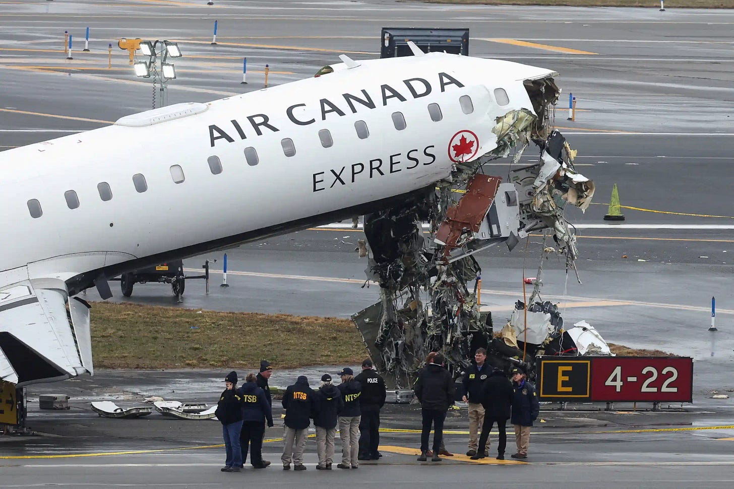 A photograph of the crashed Air Canada CRJ900 on the runway at Laguardia Airport. The front part of the airplane is visible, canted upwards. The cockpit is crushed and a tangle of debris hangs from the front of the fuselage. The words “Air Canada Express” are visible on the side of the plane, as is the red Air Canada maple leaf emblem. A group of about ten accident inspectors are gathered in front of the airplane. They wear dark jackets labeled “NTSB” in yellow or white letters. Below the nose of the aircraft, a taxiway sign is visible. It reads, E 4-22.