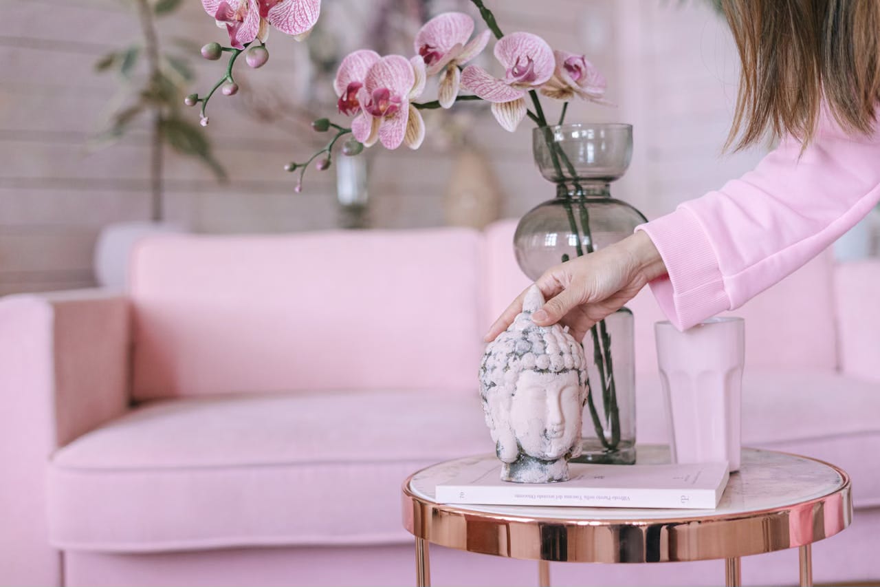 A woman places a statue of the buddha on a table next to a pink sofa and pink orchids.